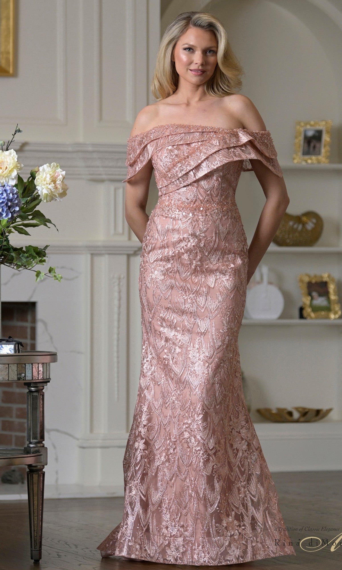A woman in the Rina Di Montella RD3531 long formal dress in rose stands indoors near a white bookshelf and flower arrangement—an ideal mother-of-the-bride look.